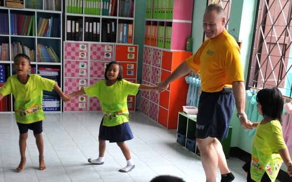 An adult male wearing a yellow shirt with 'NAVY' text is engaging in a circle game or dance activity with a group of young children in what appears to be a colorful classroom or kindergarten setting. The children are dressed in bright green school uniforms and pink casual clothing, holding hands and moving together. The room is well-equipped with bookshelves, colorful storage lockers, and educational materials on the floor.