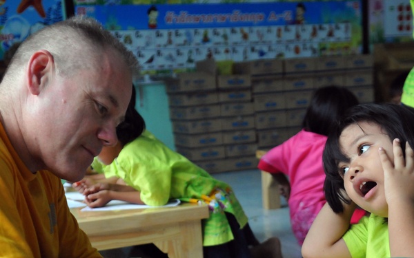 An adult male in an orange shirt is interacting with a young child in a green uniform at a classroom table. The child appears engaged and expressive, looking up at the adult with an animated facial expression. The setting appears to be an early childhood classroom, likely in Southeast Asia based on the Thai-language educational posters visible in the background.