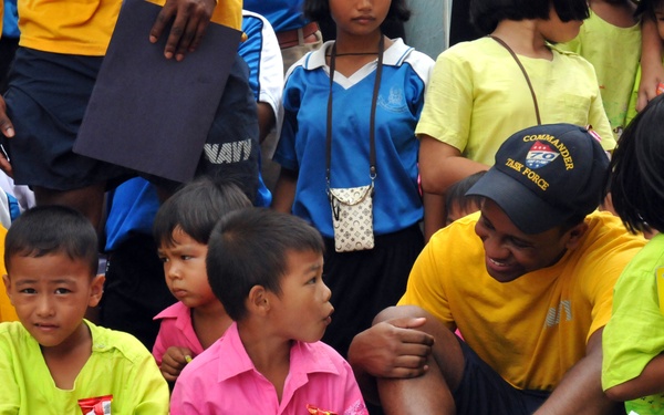 A group of children and adults are gathered together in what appears to be a community or outreach event. An adult wearing a 'Commander Task Force' cap and a yellow 'NAVY' shirt is seated among children, interacting warmly with them. The children are holding books and magazines, suggesting a reading or educational activity is taking place.