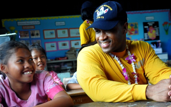 A man wearing a yellow long-sleeve shirt and a navy blue cap with 'CVN-73' (USS George Washington) insignia interacts warmly with children in what appears to be a classroom setting. He is wearing a floral lei and smiling alongside two young girls in pink shirts. The setting suggests a community outreach or humanitarian visit, likely by a U.S. Navy sailor.