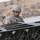 A U.S. Army soldier identified by name tape as 'BARNES' is inspecting or handling military equipment, appearing to be stacked M18A1 Claymore antipersonnel mines or similar military ordnance containers. The soldier is wearing Army Combat Uniform (ACU) in digital camouflage pattern with a matching helmet and an American flag patch on the left shoulder. The setting appears to be an outdoor military supply or staging area with additional equipment and wooden crates visible in the background.