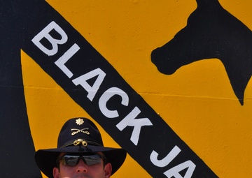 A U.S. Army soldier wearing ACU (Army Combat Uniform) stands in front of a large painted mural of the 1st Cavalry Division 'Black Jack' insignia. The soldier is identified by name tape as 'AVENT' and wears a major's oak leaf cluster insignia along with a cavalry Stetson hat. The background mural features the distinctive yellow and black shield of the 1st Cavalry Division with the text 'BLACK JACK'.