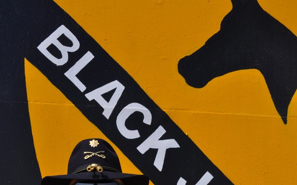 A U.S. Army soldier wearing ACU (Army Combat Uniform) stands in front of a large painted mural of the 1st Cavalry Division 'Black Jack' insignia. The soldier is identified by name tape as 'AVENT' and wears a major's oak leaf cluster insignia along with a cavalry Stetson hat. The background mural features the distinctive yellow and black shield of the 1st Cavalry Division with the text 'BLACK JACK'.