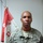 A U.S. Army soldier named Cofield poses for what appears to be an official or semi-official portrait photo. He is wearing the Army Combat Uniform (ACU) in the Universal Camouflage Pattern. A red and white unit guidon flag is visible in the background.