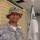 A U.S. Army soldier named Grant stands in front of a yellow brick building wearing Army Combat Uniform (ACU) in digital camouflage pattern. The soldier is wearing a boonie hat with a rank insignia, orange-tinted protective eyewear, and has a name tape reading 'GRANT' and a 'U.S. ARMY' tape visible on the uniform. A military unit guidon flag is visible in the background.