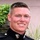 A young male in a dark military dress uniform is smiling at the camera in what appears to be an outdoor setting. The uniform features gold/brass buttons and an Eagle, Globe, and Anchor emblem visible at the collar, consistent with United States Marine Corps dress blues. The background includes green foliage and a light-colored wall.