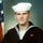 A formal military portrait of a young male U.S. Navy enlisted sailor wearing the traditional dark navy dress uniform (crackerjack) with white dixie cup hat. An American flag is partially visible in the background to the left. The photo appears to be a standard military service portrait, likely taken at boot camp graduation or early in the subject's naval career.
