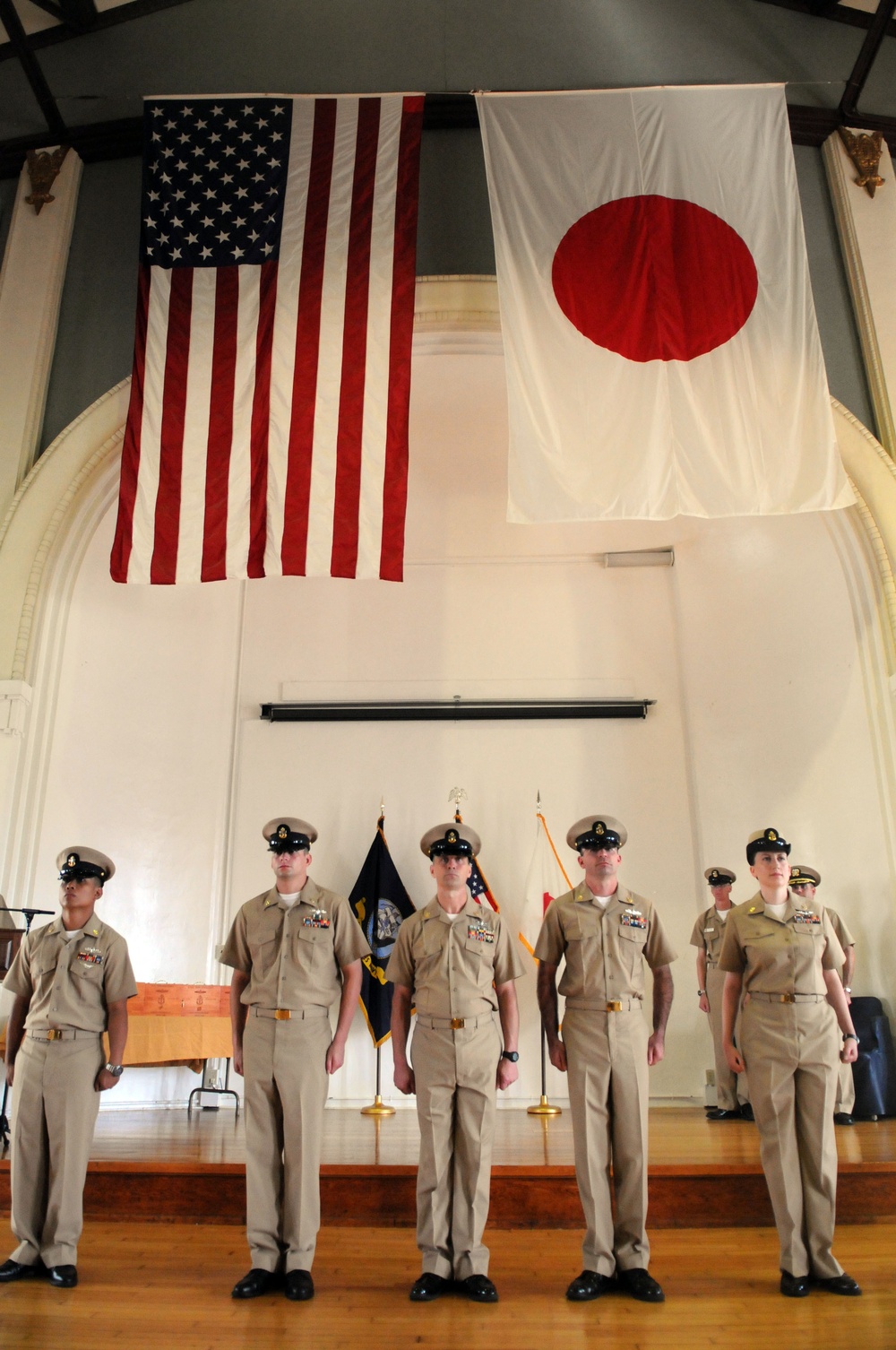 A formal military ceremony is taking place in a hall with both the United States flag and the Japanese flag (Hinomaru) prominently displayed on the wall. Five U.S. Navy personnel in khaki service uniforms stand at attention on a raised wooden stage, with additional personnel visible in the background. The setting suggests a joint U.S.-Japan military event or ceremony.