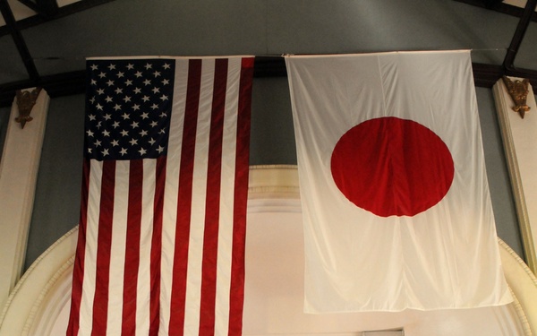 A formal military ceremony is taking place in a hall with both the United States flag and the Japanese flag (Hinomaru) prominently displayed on the wall. Five U.S. Navy personnel in khaki service uniforms stand at attention on a raised wooden stage, with additional personnel visible in the background. The setting suggests a joint U.S.-Japan military event or ceremony.