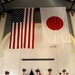 A formal military ceremony is taking place in a hall with both the United States flag and the Japanese flag (Hinomaru) prominently displayed on the wall. Five U.S. Navy personnel in khaki service uniforms stand at attention on a raised wooden stage, with additional personnel visible in the background. The setting suggests a joint U.S.-Japan military event or ceremony.