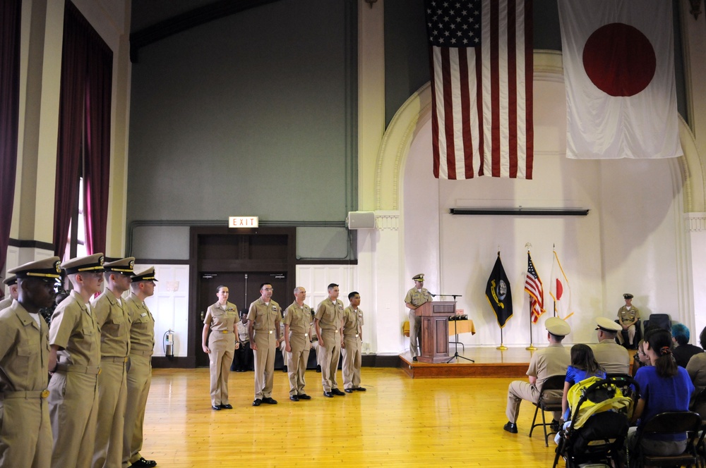 A formal U.S. Navy military ceremony is taking place in a large hall, with personnel in tan/khaki service uniforms standing at attention or in formation. Both the American flag and the Japanese flag are prominently displayed on the wall, indicating this event likely takes place at a U.S. naval installation in Japan. A speaker at a podium addresses the assembled personnel while civilians and additional military members observe from seated positions.