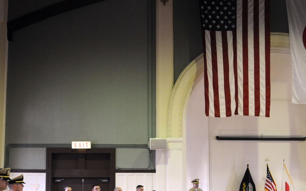 A formal U.S. Navy military ceremony is taking place in a large hall, with personnel in tan/khaki service uniforms standing at attention or in formation. Both the American flag and the Japanese flag are prominently displayed on the wall, indicating this event likely takes place at a U.S. naval installation in Japan. A speaker at a podium addresses the assembled personnel while civilians and additional military members observe from seated positions.