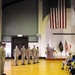 A formal U.S. Navy military ceremony is taking place in a large hall, with personnel in tan/khaki service uniforms standing at attention or in formation. Both the American flag and the Japanese flag are prominently displayed on the wall, indicating this event likely takes place at a U.S. naval installation in Japan. A speaker at a podium addresses the assembled personnel while civilians and additional military members observe from seated positions.