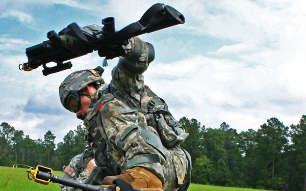 Three U.S. Army soldiers in ACU (Army Combat Uniform) camouflage are conducting what appears to be a training exercise at an outdoor range. They are operating what appears to be a Javelin or similar man-portable anti-tank missile system mounted on a wooden sawhorse-style stand. The setting is a grassy field with tree line in the background under a partly cloudy sky.