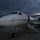 New SACEUR makes first visit to Kosovo A white private business jet is parked on an airport tarmac with its boarding stairs deployed, photographed from a low front-quarter angle. The aircraft features a gold accent stripe along the fuselage and a dark red/maroon color band near the cabin windows. The scene is captured under dramatic, overcast storm clouds at dusk or dawn, with a hangar visible in the background.