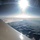 New SACEUR makes first visit to Kosovo An aerial photograph taken from inside a commercial aircraft, showing the view over a vast cloudscape from high altitude. The aircraft wing and winglet are visible in the lower-left foreground, with scattered cumulus clouds below and a bright sun causing significant lens flare in the upper portion of the frame. The horizon curves slightly in the distance, with hazy atmospheric layers visible.