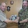 New SACEUR makes first visit to Kosovo Two military officers are shaking hands while jointly holding a commemorative plaque or shield. The officer on the left wears a US Navy tan uniform with what appear to be admiral stars, while the officer on the right wears Italian Army camouflage with a NATO patch and a name tape reading 'GAY'. The setting appears to be a military office with unit plaques and insignia mounted on a wood-paneled wall.