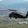 New SACEUR makes first visit to Kosovo A black military helicopter marked 'KFOR' is captured in flight over a rural town or village, with rolling hills and mountains visible in the background. The helicopter appears to be a UH-60 Black Hawk type aircraft operating under NATO's Kosovo Force (KFOR) mission. The image is taken from another airborne platform, providing an aerial perspective of both the aircraft and the landscape below.
