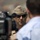 New SACEUR makes first visit to Kosovo A U.S. Marine Corps officer wearing a garrison cover (cap) with four silver stars and the Eagle, Globe, and Anchor emblem is being interviewed or filmed outdoors. The officer is wearing sunglasses and a tan/khaki uniform, facing a civilian in a light blue shirt while a video camera is visible in the foreground. The background is blurred, suggesting a public or official outdoor event.