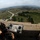 New SACEUR makes first visit to Kosovo Military personnel are gathered on top of an elevated observation post or watchtower, overlooking a wide rural landscape with a town, industrial facilities, and open fields visible in the distance. Several soldiers in different uniforms are examining what appears to be maps or documents on a flat surface, alongside binoculars. The image is captured with a fisheye lens, creating a wide-angle distorted perspective.
