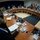 New SACEUR makes first visit to Kosovo A multinational military conference or briefing is taking place in a formal meeting room arranged in a horseshoe/U-shaped table configuration. Numerous military personnel in various national uniforms are seated around the tables, along with at least one civilian in a dark suit in the foreground. Multiple national flags are displayed along the back wall, indicating an international or coalition military gathering.