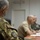 New SACEUR makes first visit to Kosovo A military briefing or meeting is taking place in an indoor conference room setting. The foreground shows a soldier wearing Italian-pattern camouflage (Vegetato) with a NATO COMKFOR (Commander Kosovo Force) patch on the sleeve. In the background, at least two other military personnel are seated at a table with microphones and documents.