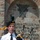 New SACEUR makes first visit to Kosovo A uniformed bagpiper is playing bagpipes in front of a stone monument or memorial wall with Cyrillic script inscription. The piper wears a white short-sleeve uniform shirt with a black tie, orange/saffron kilt, white belt, and a black beret with a gold badge and green hackle. The stone wall behind features a carved relief depicting crossed swords or sabers within a shield, consistent with Serbian military or historical memorial iconography.