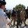 New SACEUR makes first visit to Kosovo A military ceremony or formal procession is taking place outdoors on a cobblestone plaza, likely at a military installation or memorial site. A bagpiper in a white shirt with military patches stands at attention on the left, while a group of multinational military personnel and at least one civilian walk past. The personnel are wearing various camouflage uniforms and berets, suggesting a NATO or multinational military event.