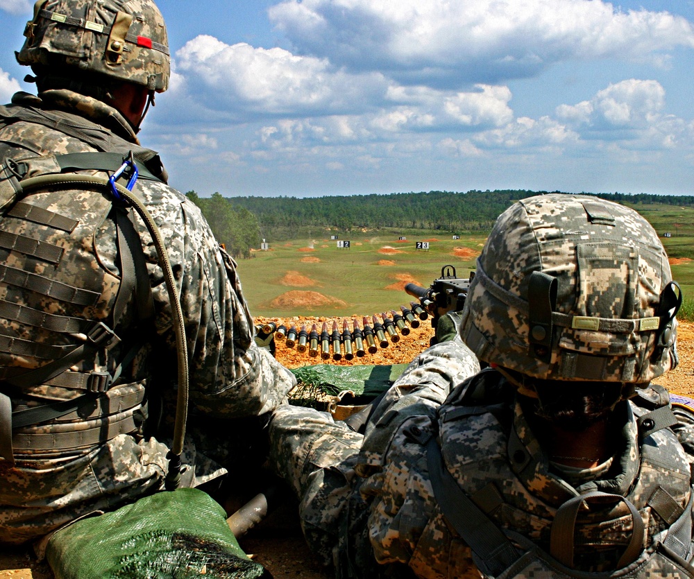 Two U.S. Army soldiers in ACU (Army Combat Uniform) digital camouflage are positioned at what appears to be a live-fire training range. One soldier is operating or preparing to operate a belt-fed machine gun, with a visible ammunition belt loaded with rounds. Numbered target markers are visible downrange across an open field bordered by treeline.