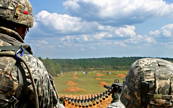Two U.S. Army soldiers in ACU (Army Combat Uniform) digital camouflage are positioned at what appears to be a live-fire training range. One soldier is operating or preparing to operate a belt-fed machine gun, with a visible ammunition belt loaded with rounds. Numbered target markers are visible downrange across an open field bordered by treeline.
