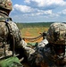 Two U.S. Army soldiers in ACU (Army Combat Uniform) digital camouflage are positioned at what appears to be a live-fire training range. One soldier is operating or preparing to operate a belt-fed machine gun, with a visible ammunition belt loaded with rounds. Numbered target markers are visible downrange across an open field bordered by treeline.