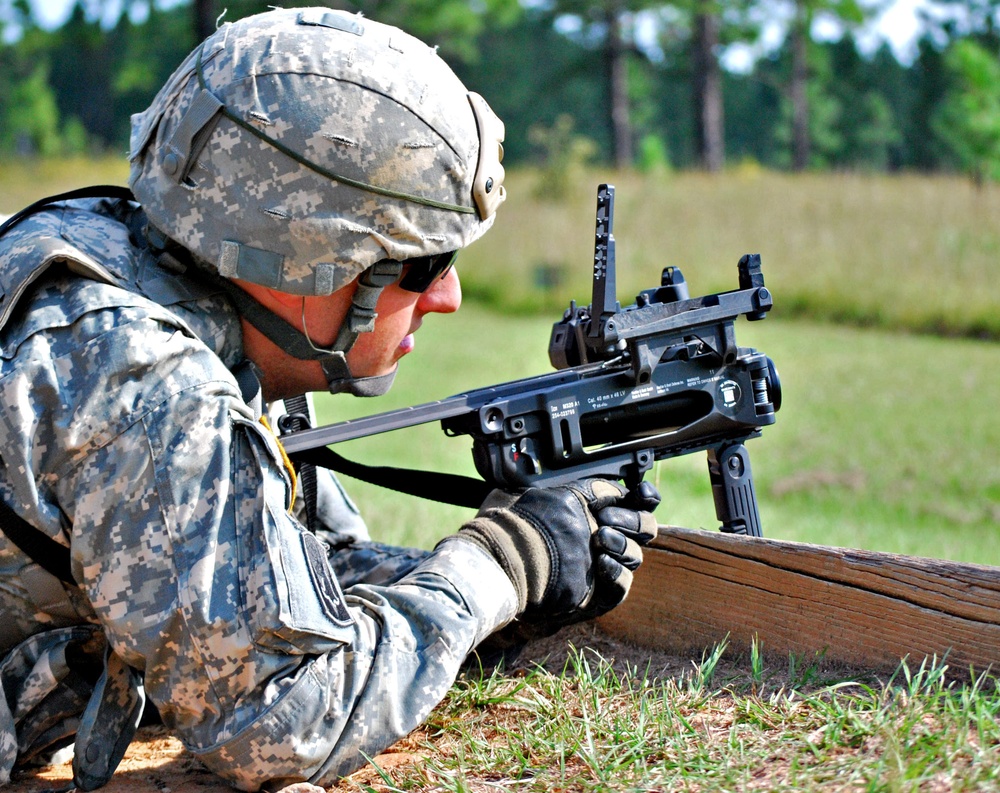 A U.S. Army soldier in full combat gear is in a prone firing position at what appears to be a military training range. The soldier is aiming an M320 grenade launcher (standalone configuration) over a wooden barrier. The setting is an outdoor range with green grass and trees visible in the background.