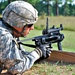 A U.S. Army soldier in full combat gear is in a prone firing position at what appears to be a military training range. The soldier is aiming an M320 grenade launcher (standalone configuration) over a wooden barrier. The setting is an outdoor range with green grass and trees visible in the background.