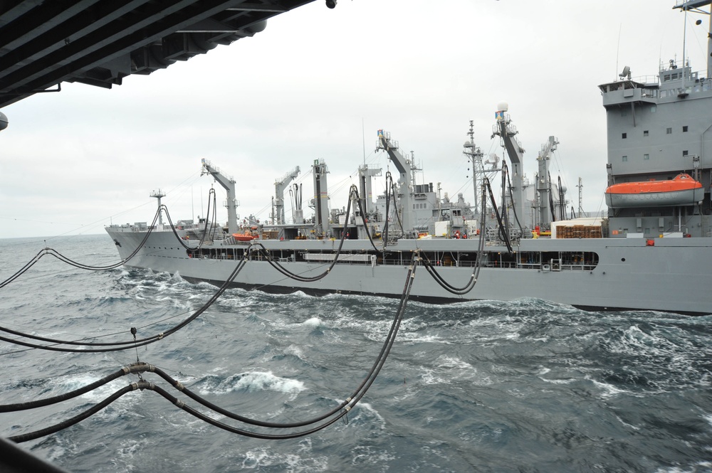 The image depicts a replenishment at sea (RAS) operation between two naval vessels. Multiple fuel/transfer hoses are strung between the ships while underway on choppy, overcast seas. The supply vessel in the center is equipped with multiple replenishment rigs and cranes, indicating a combat stores or fleet replenishment oiler.