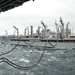 The image depicts a replenishment at sea (RAS) operation between two naval vessels. Multiple fuel/transfer hoses are strung between the ships while underway on choppy, overcast seas. The supply vessel in the center is equipped with multiple replenishment rigs and cranes, indicating a combat stores or fleet replenishment oiler.