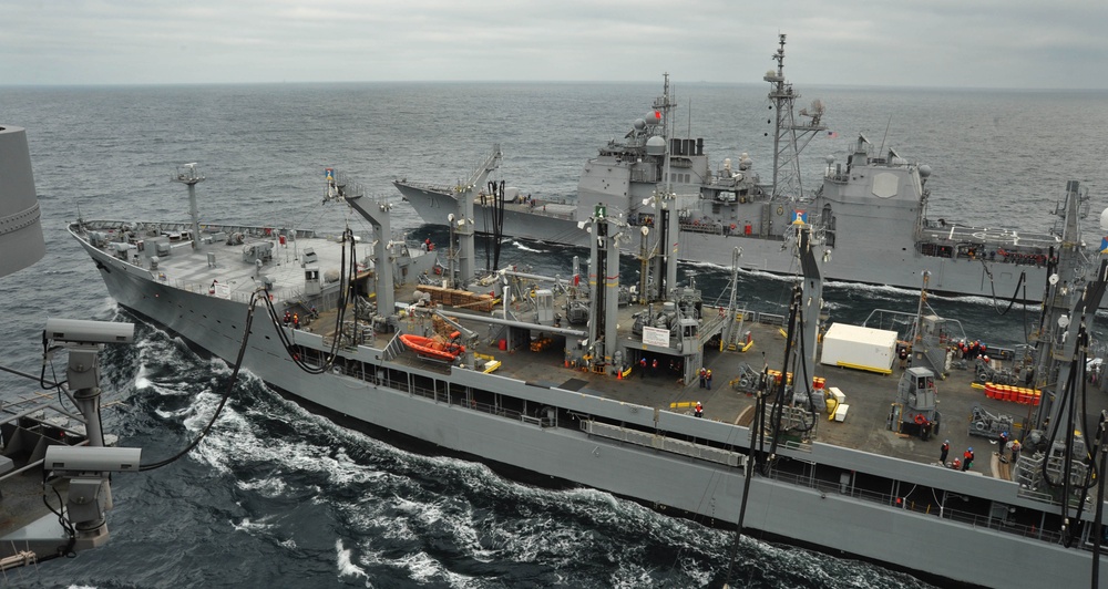 The image depicts a replenishment-at-sea (RAS) operation involving multiple U.S. Navy vessels underway in open ocean conditions. A large auxiliary replenishment ship is positioned centrally, connected to at least two warships on either side via transfer rigs and lines. The operation appears to be actively transferring fuel and/or supplies while all vessels maintain parallel courses.