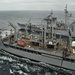 The image depicts a replenishment-at-sea (RAS) operation involving multiple U.S. Navy vessels underway in open ocean conditions. A large auxiliary replenishment ship is positioned centrally, connected to at least two warships on either side via transfer rigs and lines. The operation appears to be actively transferring fuel and/or supplies while all vessels maintain parallel courses.