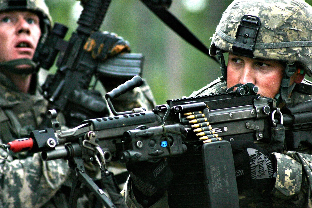 Two soldiers in U.S. Army ACU (Army Combat Uniform) digital camouflage are shown in what appears to be a field training exercise. The foreground soldier is aiming an M249 Squad Automatic Weapon (SAW) with a visible ammunition belt loaded into the weapon. Both soldiers are wearing standard-issue ACU helmets and tactical gear.