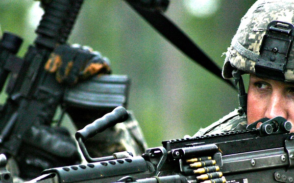 Two soldiers in U.S. Army ACU (Army Combat Uniform) digital camouflage are shown in what appears to be a field training exercise. The foreground soldier is aiming an M249 Squad Automatic Weapon (SAW) with a visible ammunition belt loaded into the weapon. Both soldiers are wearing standard-issue ACU helmets and tactical gear.