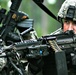 Two soldiers in U.S. Army ACU (Army Combat Uniform) digital camouflage are shown in what appears to be a field training exercise. The foreground soldier is aiming an M249 Squad Automatic Weapon (SAW) with a visible ammunition belt loaded into the weapon. Both soldiers are wearing standard-issue ACU helmets and tactical gear.