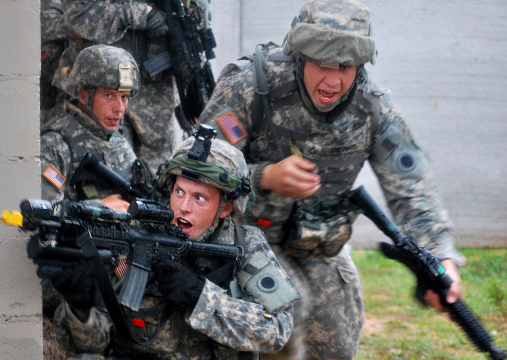 The image shows U.S. Army soldiers in Army Combat Uniform (ACU) conducting what appears to be a military training exercise in an urban environment. Multiple soldiers are taking cover at a building corner, with at least one aiming an M4 carbine rifle while another appears to be issuing commands. The expressions and body language suggest a dynamic, high-intensity training scenario.