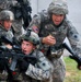 The image shows U.S. Army soldiers in Army Combat Uniform (ACU) conducting what appears to be a military training exercise in an urban environment. Multiple soldiers are taking cover at a building corner, with at least one aiming an M4 carbine rifle while another appears to be issuing commands. The expressions and body language suggest a dynamic, high-intensity training scenario.