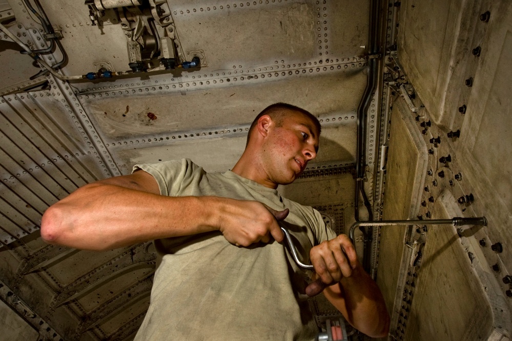 A young male technician in a olive/tan t-shirt is working inside what appears to be the interior of a large military aircraft, likely performing maintenance. He is using a hand tool, possibly a wrench or ratchet, while working among visible pipes, conduits, riveted metal panels, and mechanical components. The photo is taken from a low angle, looking upward, giving a dramatic perspective of the confined workspace.