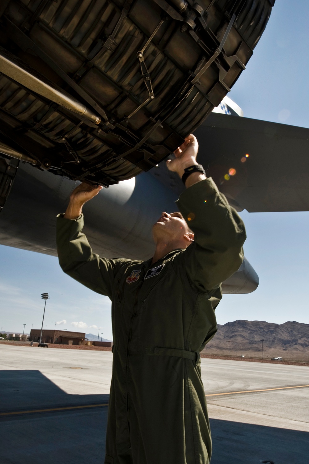 A U.S. Air Force personnel wearing an olive green flight suit is conducting a pre-flight or post-flight inspection of a large military aircraft engine nacelle on a flight line. The individual is reaching upward to examine the underside of what appears to be a turbofan engine. The setting appears to be a desert air base, consistent with locations such as Nellis AFB or a similar southwestern U.S. installation.