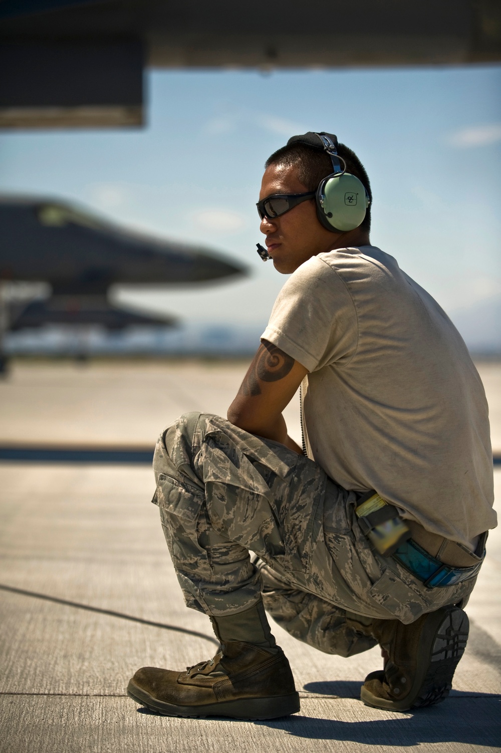 A military aircraft maintainer or crew chief is crouching on an airfield tarmac, wearing hearing protection headset and safety goggles. He is dressed in US Air Force ABU (Airman Battle Uniform) camouflage pants and a tan t-shirt, with a tattoo visible on his upper arm. A military jet aircraft is visible in the blurred background on the flight line.