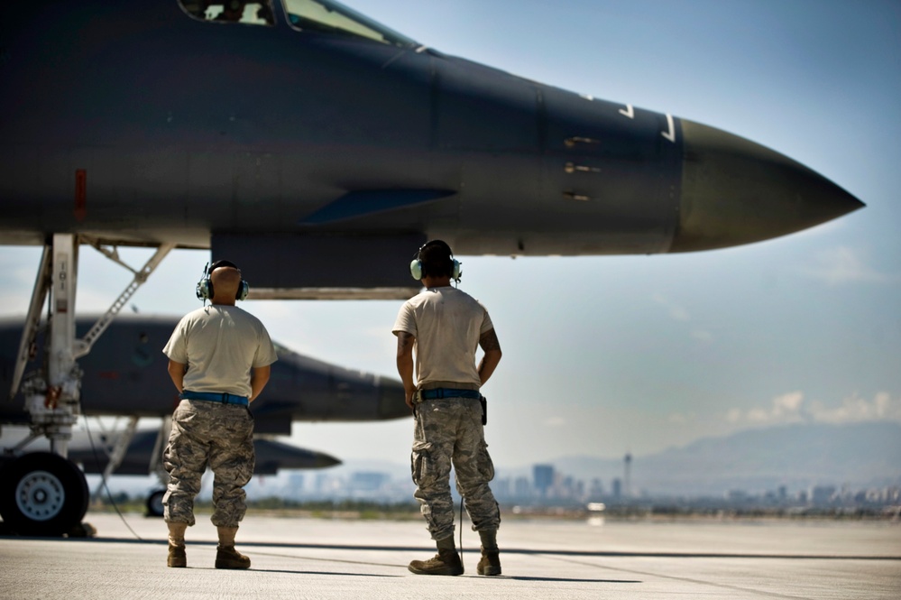 Two U.S. Air Force ground crew personnel in ABU (Airman Battle Uniform) stand on a flight line observing a fighter jet, likely an F-16 Fighting Falcon, during what appears to be pre-flight or post-flight operations. Both airmen are wearing hearing protection headsets and have blue belts, indicating crew chief or maintenance roles. A city skyline and mountain range are visible in the background, consistent with Nellis Air Force Base near Las Vegas, Nevada.