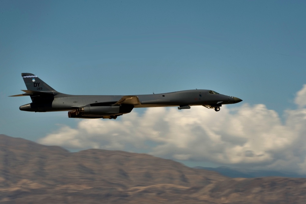 A U.S. Air Force B-1B Lancer strategic bomber is captured in low-altitude flight, likely during takeoff or a low-level pass. The aircraft bears the tail code 'DY' and a checkered tail stripe, identifying it as assigned to Dyess Air Force Base, Texas. Arid mountainous terrain and partial cloud cover form the backdrop.