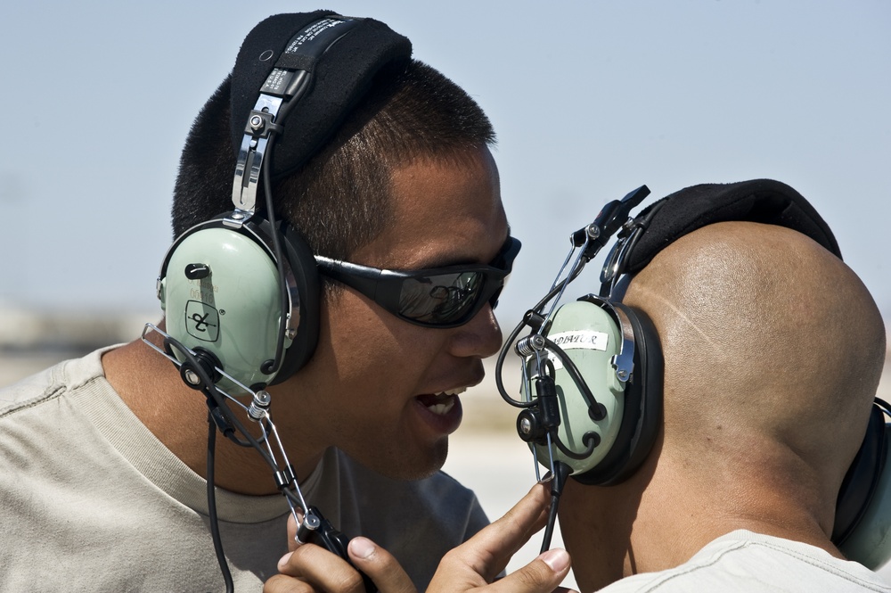 Two individuals wearing military-style aviation headsets are engaged in close communication outdoors, likely on a flight line or airfield. The person facing the camera is wearing dark wraparound sunglasses and appears to be speaking directly to the other individual. Both are wearing tan/beige shirts consistent with military utility uniforms.
