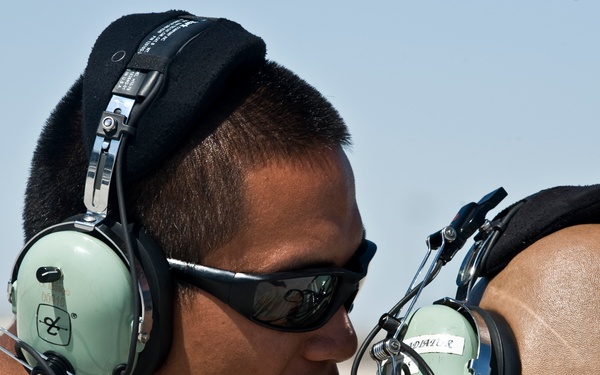 Two individuals wearing military-style aviation headsets are engaged in close communication outdoors, likely on a flight line or airfield. The person facing the camera is wearing dark wraparound sunglasses and appears to be speaking directly to the other individual. Both are wearing tan/beige shirts consistent with military utility uniforms.