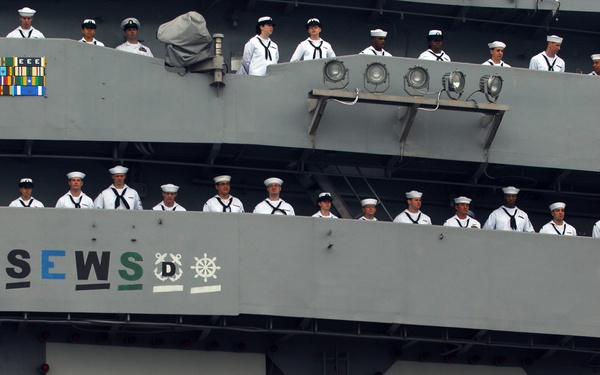 The image shows the hull and superstructure of a US Navy aircraft carrier bearing the hull number 73, identified as USS George Washington (CVN-73). Numerous sailors in white dress uniforms are manning the rails across multiple deck levels, a formal naval tradition typically performed during port arrivals or departures. The large illuminated hull numbers '73' dominate the lower portion of the image, surrounded by navigation lights.